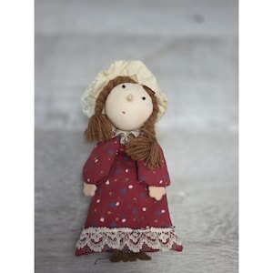 May include: A handmade rag doll with brown yarn hair in braids, wearing a red floral dress with a white lace hem and a cream-colored bonnet. The doll has a stitched face with small black eyes and a red mouth.