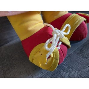May include: Close-up of a doll's feet wearing red and yellow shoes with white laces. The shoes have yellow trim and are tied with a white shoelace bow. The doll's feet are made of red fabric.