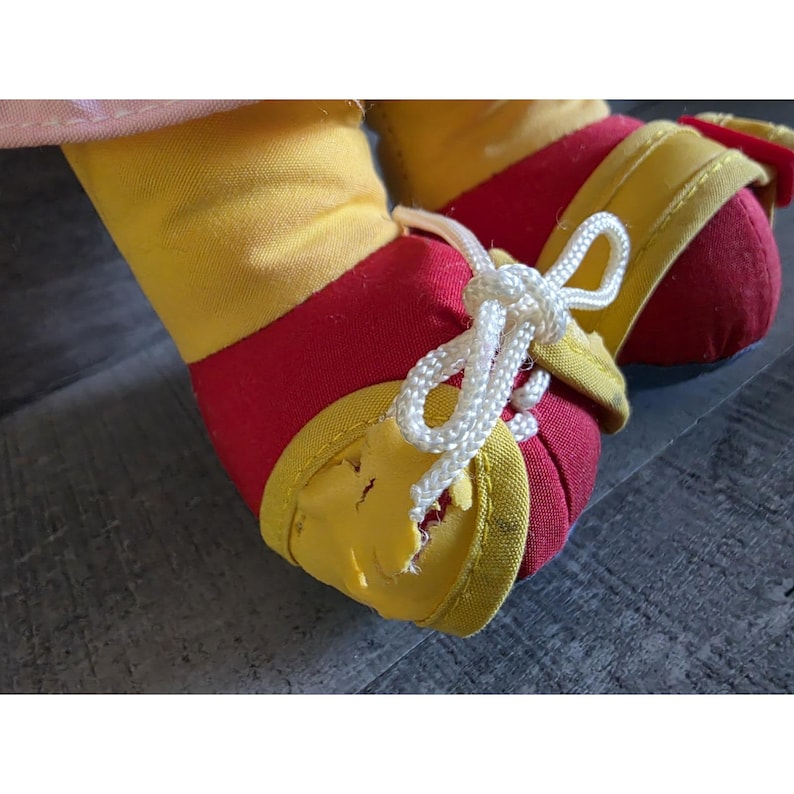 May include: Close-up of a doll's feet wearing red and yellow shoes with white laces. The shoes have yellow trim and are tied with a white shoelace bow. The doll's feet are made of red fabric.