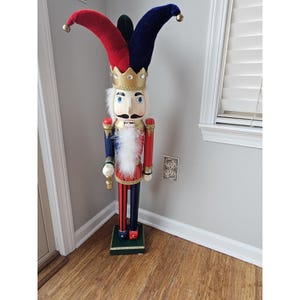 May include: A wooden nutcracker with a red and blue jester hat, a white beard, and a gold crown. The nutcracker is holding a golden staff in its right hand.