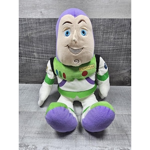 May include: A soft toy of Buzz Lightyear from Toy Story. The doll has a purple head, a white and green spacesuit, and purple feet. The word "LIGHTYEAR" is printed on the chest.