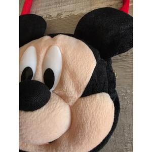 May include: A plush, black, and tan Mickey Mouse-shaped bag with large white eyes and a red strap. The bag features a zipper closure and is made of soft, textured fabric. The design is a close-up of Mickey's face.