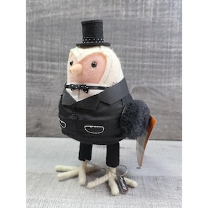 May include: A whimsical owl figurine dressed in a black tuxedo with a matching top hat and bow tie. The owl has a cream-colored face and a peach-colored beak. Its feet are white, and it has a small tag attached.