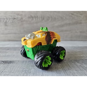 Puede incluir: Un juguete de camión monstruo amarillo con ruedas verdes y un diseño tribal marrón y naranja en el lateral.