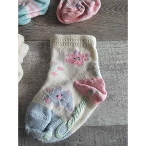 Puede incluir: Un par de calcetines blancos para beb&eacute; con un estampado floral azul y rosa. Los calcetines tienen la palabra "Carter's" escrita en verde en la parte inferior.