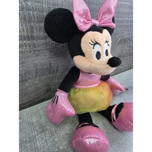 K&ouml;nnte beinhalten: Eine Pl&uuml;schpuppe von Minnie Mouse mit schwarzem Kopf und Ohren, einer rosa Schleife und einem rosa und gelben Kleid. Die Puppe hat schwarze Arme und Beine sowie rosa Handschuhe und Schuhe. Die Puppe sitzt auf einer grauen Oberfl&auml;che.