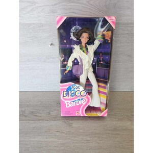 Pode incluir: Uma boneca vintage Barbie dos anos 70, "Disco Barbie", vestida com um macacão branco e uma bolsa roxa. A boneca está em pé, com um braço levantado, em uma pista de dança rosa e branca. A caixa é rosa e branca com o texto "70s Disco Barbie" em rosa e branco.