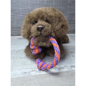 Op de afbeelding: Een bruin pluche hondenspeeltje met een touw in zijn bek. Het touw is een gedraaid ontwerp van paars en oranje. De hond heeft een pluizig uiterlijk en zit rechtop. Het speelgoed is ontworpen voor huisdieren.