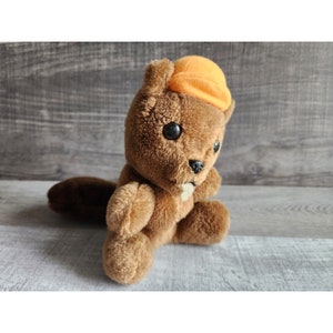 Puede incluir: Un juguete de peluche de castor marrón que lleva un sombrero naranja. El castor tiene un pequeño diente blanco que sobresale de su boca.