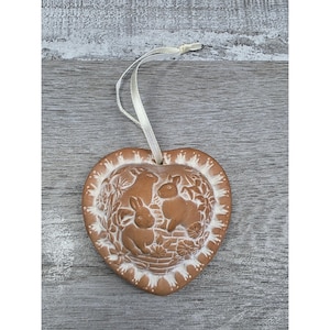Peut inclure: Ornement en céramique marron en forme de cœur, avec un motif en relief de lapins et de feuillage. Il a une bordure blanche et un ruban crème pour l'accrocher. Le motif comprend deux lapins et des éléments floraux.