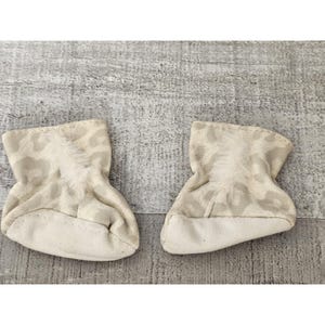 May include: A pair of white baby booties with a light brown leopard print. The booties have a soft, fuzzy texture and are perfect for keeping little feet warm.