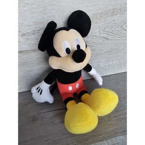 Puede incluir: Un juguete de peluche Mickey Mouse negro y amarillo sentado en una superficie de madera. El juguete lleva un atuendo rojo y blanco con detalles negros.