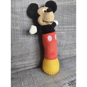 May include: A Mickey Mouse rattle toy made of plush fabric. The toy has a black head with large ears, a tan face, and white hands. The body is red with white circles and a yellow patterned base.