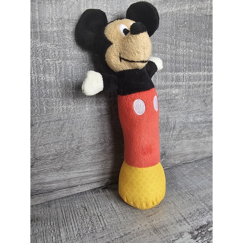 May include: A Mickey Mouse rattle toy made of plush fabric. The toy has a black head with large ears, a tan face, and white hands. The body is red with white circles and a yellow patterned base.