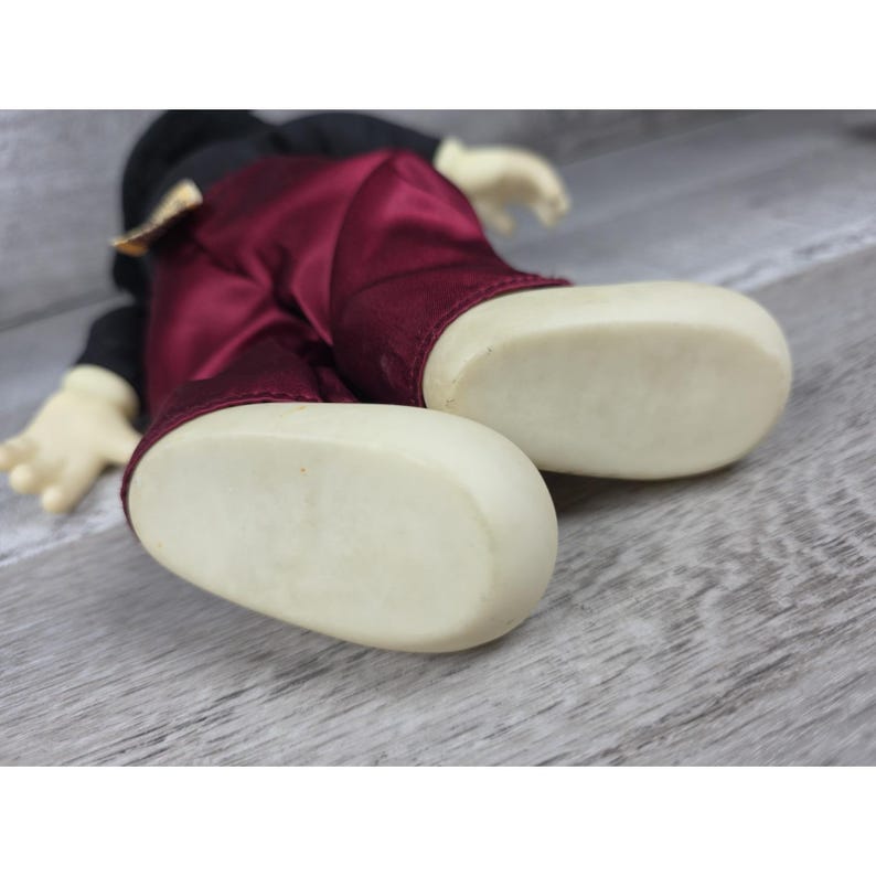 May include: A Mickey Mouse plush doll, lying down, with cream-colored feet and hands. The doll wears a black top and burgundy satin pants. The background is a light gray wooden surface.