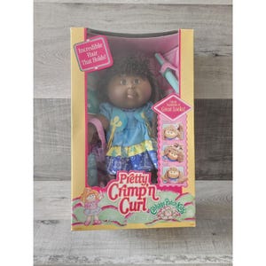 May include: A Cabbage Patch Kids doll with brown hair and a blue and yellow dress. The doll is holding a pink purse and a blue and white striped toy. The box says "Pretty Crimp'n Curl" and "Incredible Hair That Holds!"
