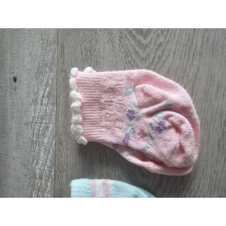 Puede incluir: Un par de calcetines de beb&eacute; de color rosa con un borde de encaje blanco y un patr&oacute;n floral. Los calcetines tienen una peque&ntilde;a inscripci&oacute;n que dice "Baby's First"