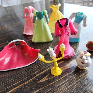 Può includere: Una collezione di personaggi giocattolo in plastica e accessori di abbigliamento per bambole che include un mantello rosso, un personaggio candelabro giallo, una piccola teiera, un pezzo di capelli marroni e vari abiti colorati in rosa, verde, giallo, blu e bianco.