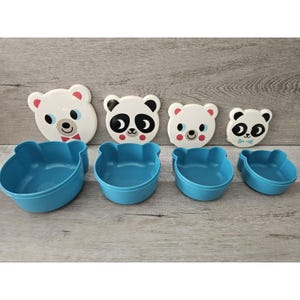 Puede incluir: Conjunto de cuatro recipientes de plástico azul con tapas blancas que presentan caras de animales de dibujos animados. Las tapas tienen un panda, un oso y dos osos polares.