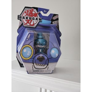 Peut inclure: Un jouet Bakugan bleu et violet avec un personnage bleu et blanc à l'intérieur. Le personnage porte un chapeau haut de forme et a un monocle. L'emballage indique "Bakugan" et "Cubbo".