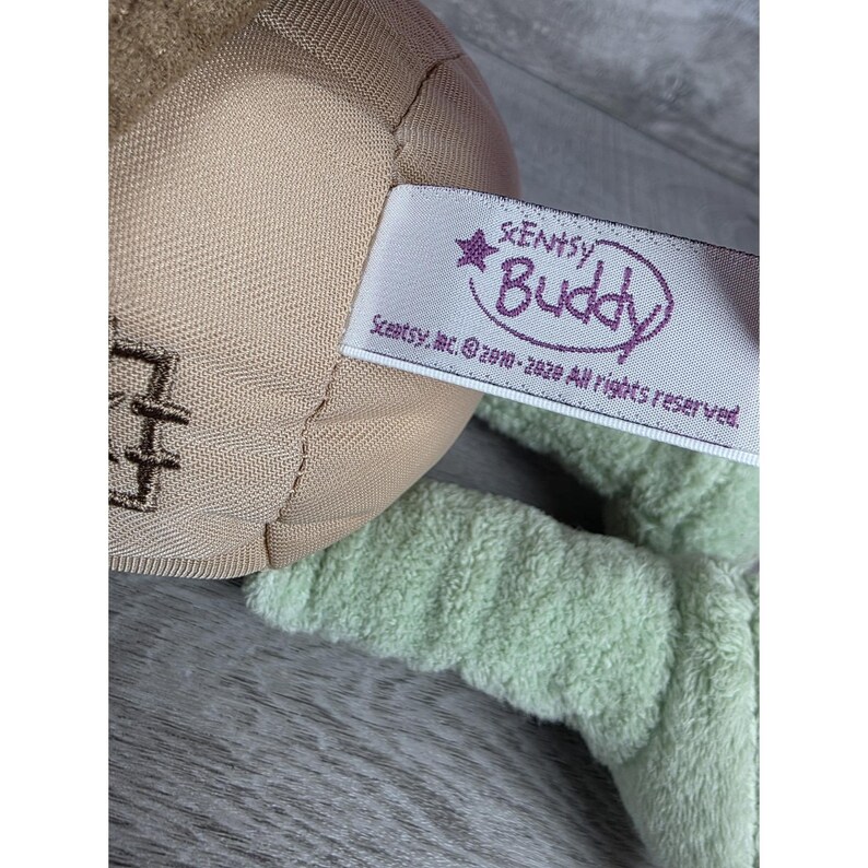 May include: Close-up of a plush toy with a light brown head and light green limbs. A white tag with the text "Scentsy Buddy" in a purple oval is attached to the toy. The tag also includes the Scentsy, Inc. copyright information.