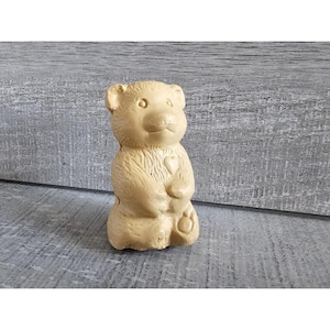 May include: A small, beige ceramic bear figurine with a heart on its chest. The bear is sitting with its paws in front of it.
