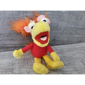 May include: A plush toy character with a yellow head, red body, and yellow feet and hands. The character has a large red mouth, orange hair, and pink accents around the eyes. The toy is sitting on a gray wooden surface.