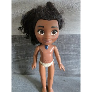 May include: A doll with dark skin, black hair, and large brown eyes. The doll is wearing a white diaper and a blue necklace. The doll's body is tan, and it has a smiling expression.