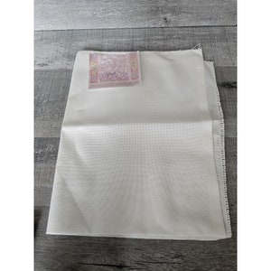 May include: A white Aida cloth with a small pink and purple label that says "DMC" on it. The cloth is folded in half and has a fringed edge.
