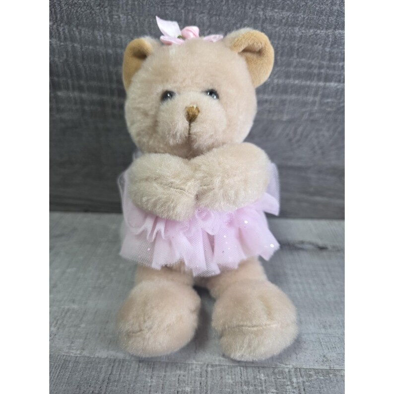 Pu&ograve; includere: Un orsacchiotto di peluche seduto, con un tut&ugrave; rosa e un fiocco abbinato. L'orso ha una pelliccia marrone chiaro con orecchie marroni pi&ugrave; scure e un naso dorato. Il tut&ugrave; &egrave; realizzato in un tessuto rosa trasparente con glitter.