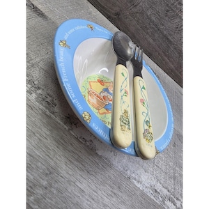 Puede incluir: Un juego de vajilla infantil que incluye un cuenco de cerámica blanco con borde azul e ilustraciones de conejos. La cuchara y el tenedor tienen mangos color crema con diseños florales y de conejos.