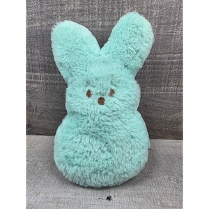 May include: A plush, light teal bunny with brown embroidered eyes and nose. The bunny has long, upright ears and a rounded body. The soft toy is designed in a classic Easter candy style.
