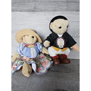 Puede incluir: Dos osos de peluche vestidos con ropa tradicional alemana. La osa lleva un sombrero de paja, un vestido azul y blanco y un lazo rojo. El oso lleva una chaqueta negra, una camisa blanca, una faja amarilla y zapatos negros. El oso tiene una etiqueta que dice "Muffi Vanderbear Santa Anna".