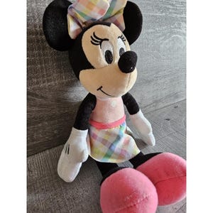 K&ouml;nnte beinhalten: Ein Pl&uuml;schtier von Minnie Mouse, das ein rosa Kleid mit einem bunten karierten Rock und einem passenden Stirnband tr&auml;gt. Das Spielzeug hat schwarze Ohren, eine schwarze Nase und rosa Schuhe.