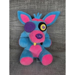 Può includere: Un peluche raffigurante un personaggio volpe stilizzato con accenti blu, rosa e viola. Il giocattolo ha grandi orecchie, un muso e un solo occhio coperto da una toppa viola. Il personaggio ha un dente giallo.