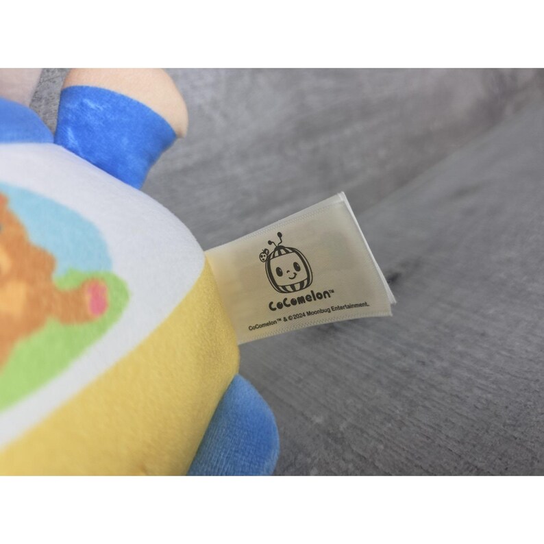 May include: A soft toy in yellow, blue, and white. The toy has a tag with the "Cocomelon" logo and text. The toy is made of fabric.