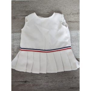 Op de afbeelding: Witte, mouwloze jurk met een geplooide rok en een rood, wit en blauw lint.