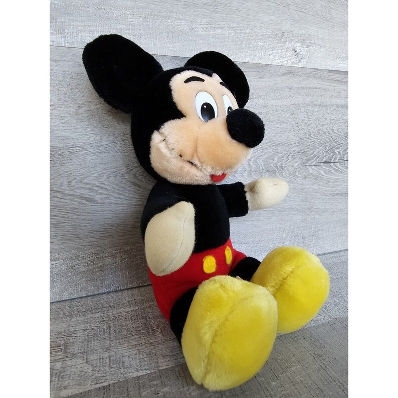 May include: A plush Mickey Mouse toy sitting with its legs crossed. The toy is black with yellow shoes and red shorts with two yellow buttons.