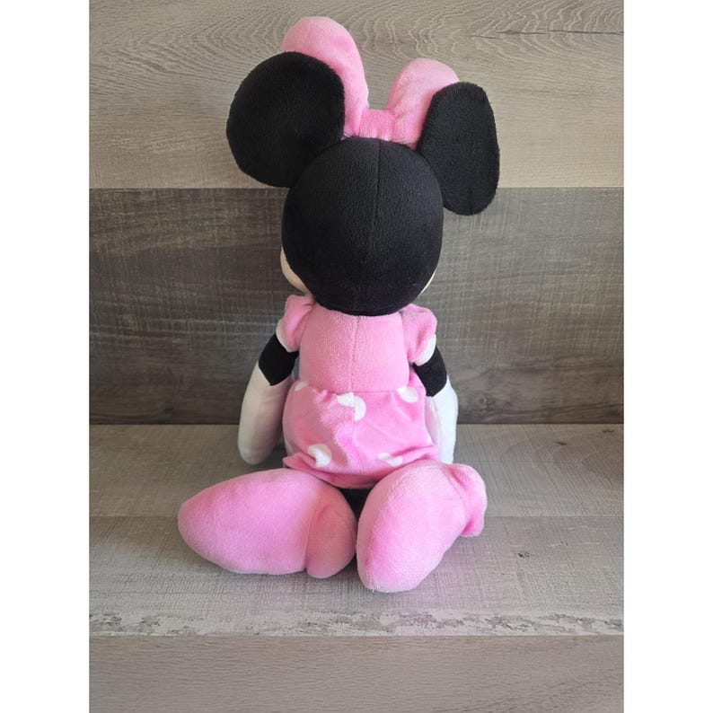 May include: A plush Minnie Mouse doll, viewed from the back, with a pink dress and bow. The doll has black ears and a black head. The dress has white polka dots. The doll is sitting on a wooden surface.