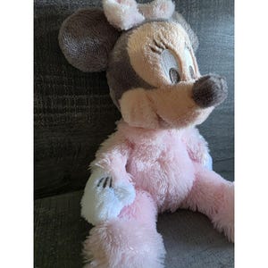 May include: A plush Minnie Mouse doll in pink, gray, and white. The doll has large, round ears, a soft pink body, and white gloves. The doll's face features embroidered details and a friendly expression.