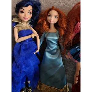 Op de afbeelding: Er worden drie poppen getoond, elk met gedetailleerde gelaatstrekken en uitgebreide kostuums. De pop links heeft blauw haar en een blauwe jurk. De middelste pop heeft rood haar en een blauwe jurk. De derde pop is gedeeltelijk zichtbaar.