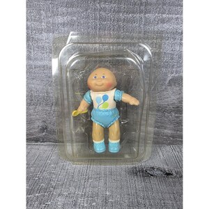 May include: A small plastic doll with a round head, rosy cheeks, and purple eyes, wearing a white top with blue and green balloon graphics and light blue ruffled shorts. The doll has light blue shoes and holds a small yellow accessory in one hand.