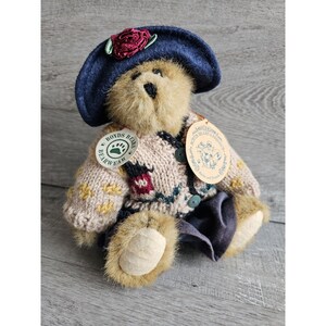 May include: A brown teddy bear wearing a blue hat with a red rose, a beige knitted sweater with floral and leaf patterns, and blue pants. The bear is sitting on a wooden surface. The bear has two tags attached to its sweater, one that says "Boyds Bears Bearwear" and another that says "Boyds Bears & Friends".