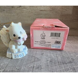 Peut inclure: Une figurine en porcelaine blanche représentant un renard portant une écharpe rose, assis sur une base bleu et blanche. La figurine est datée de 2003 et fait partie de la collection Precious Moments. "Bright Eyed & Bushy Tailed" est écrit sur la boîte.