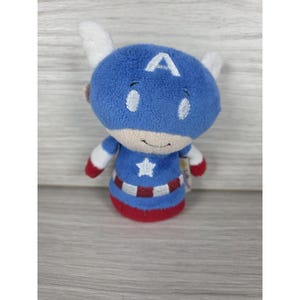 May include: A plush toy of Captain America featuring a blue head with a white letter A, white wings on the sides, and a blue body with a white star on the chest. The base of the toy has a red and white striped pattern with a solid red bottom edge.