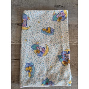 Puede incluir: Una manta para bebé de color azul claro con un patrón repetido de personajes de dibujos animados en lunas crecientes y en camas. La manta tiene un fondo floral azul claro y estrellas amarillas.