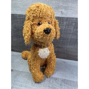 May include: A plush toy dog with curly, light brown fur. The dog has brown eyes, a black nose, and a brown collar with a white patch on its chest. The dog is sitting on a gray surface.