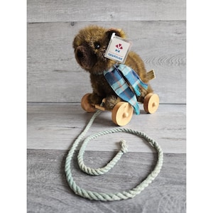 May include: A brown plush bear with a blue and white plaid scarf is attached to a wooden pull toy with wheels. The toy has a rope attached to it. The bear has a tag that says "Applause, Learning Toys That Grow".