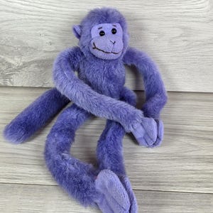May include: A plush monkey toy covered in soft purple fur. The stuffed animal features long, dangling arms and legs, a textured face with a stitched brown mouth, and small round ears. The monkey has a long tail and dark plastic eyes.