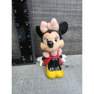 May include: A Minnie Mouse figurine with a pink bow, black ears, and a red dress. The figurine has yellow shoes and is approximately 3 inches tall.